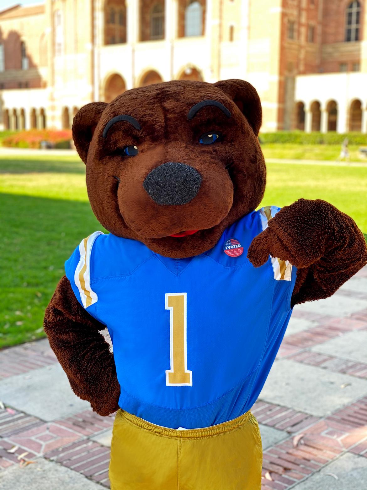 Joe Bruin (Mascot) is wearing a Blue and Yellow sports jersey standing in front of Royce Hall, pointing at a "Voter" button on their top right shirt. 